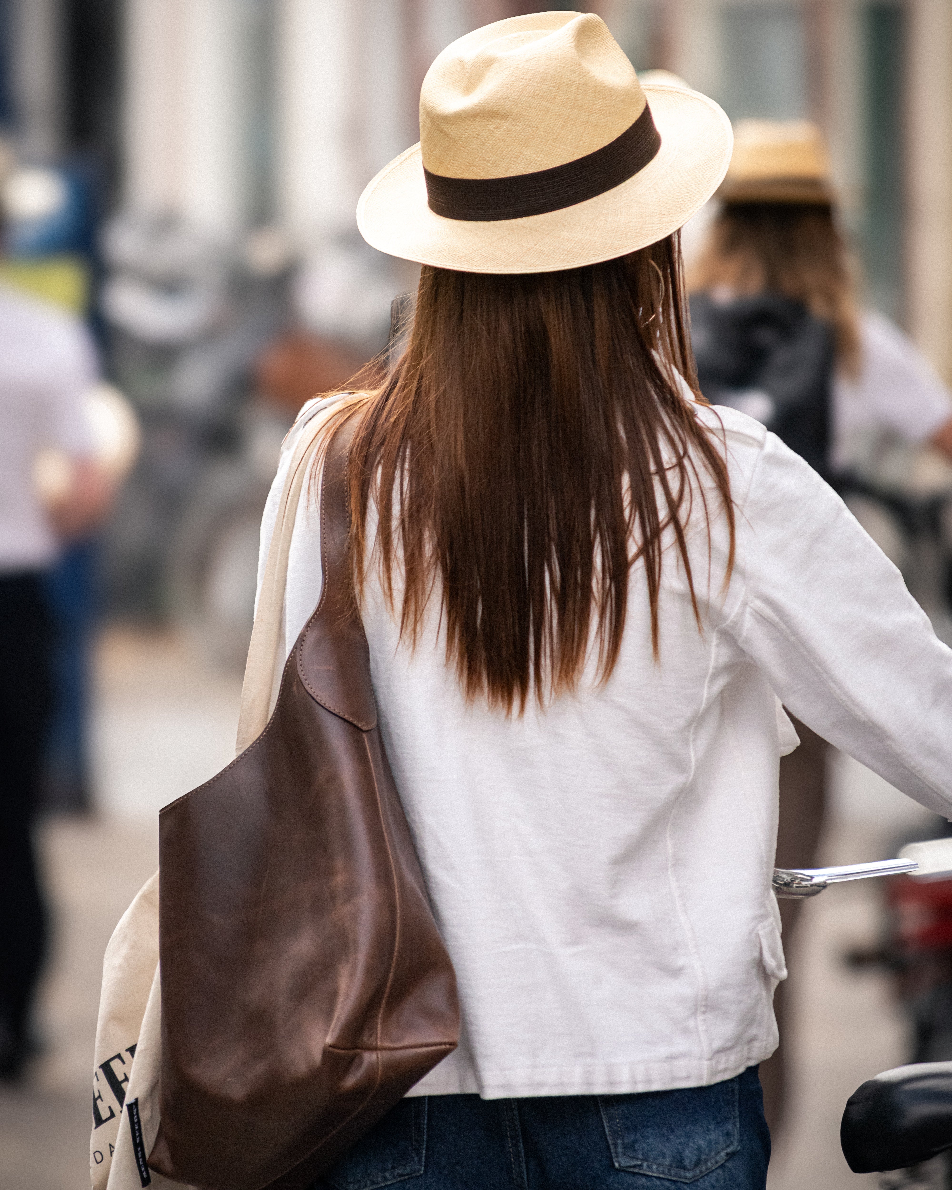 A woman seen from behind wearing a light beige fedora hat with a dark brown band, a white shirt, and blue jeans, carrying a large brown leather shoulder bag on a city street.
